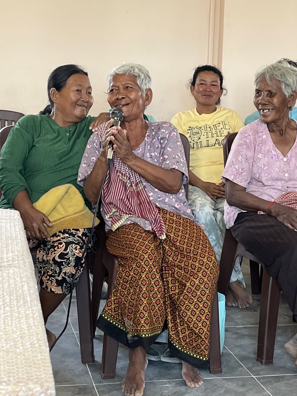 Older women from the Pursat community seated together, one singing into a microphone while the others smile