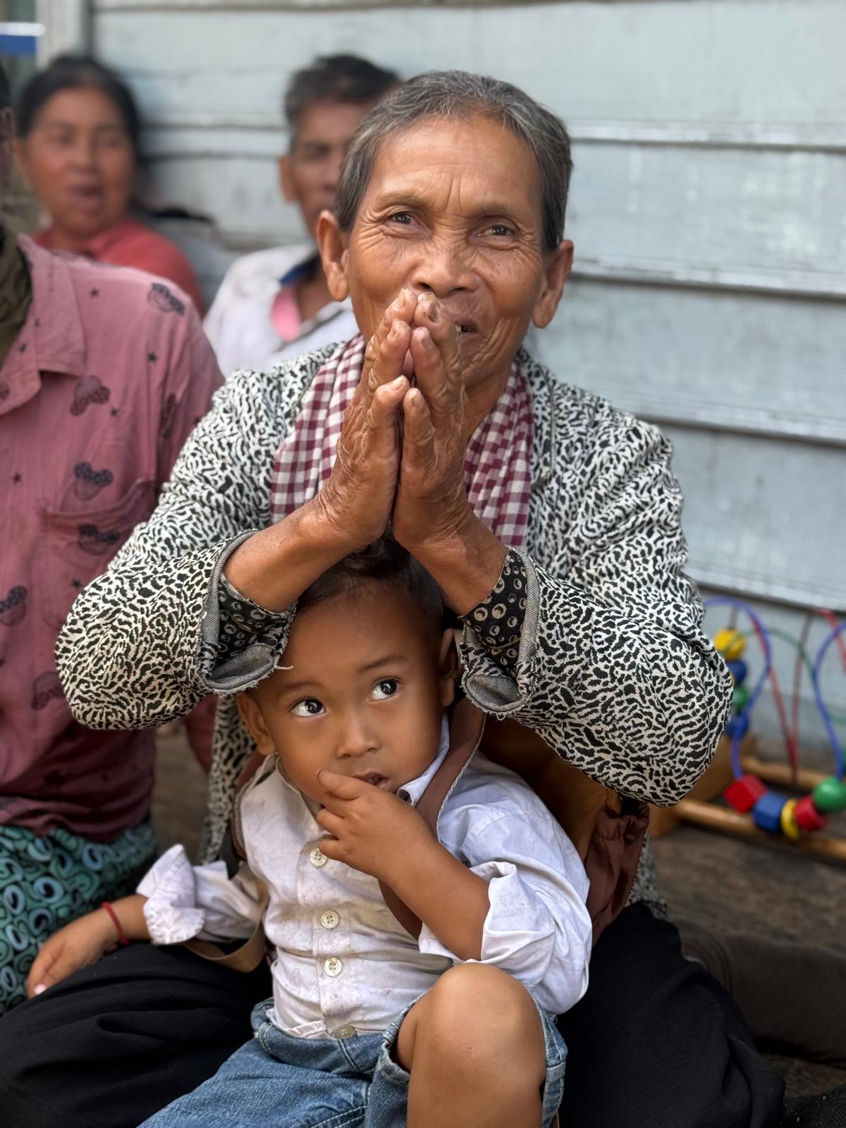 An older woman with her hands pressed together in prayer, a small child resting on her lap