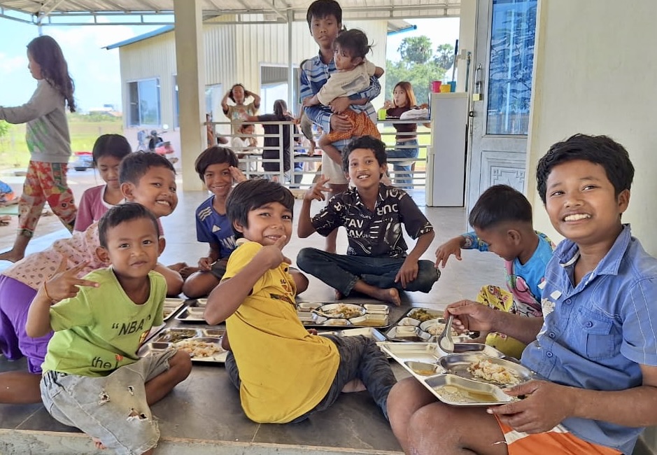 Children sitting together on the floor sharing a meal from metal trays, smiling at the camera