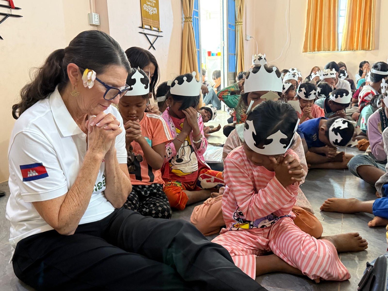 A team member kneeling in prayer beside children wearing paper crowns during a lesson