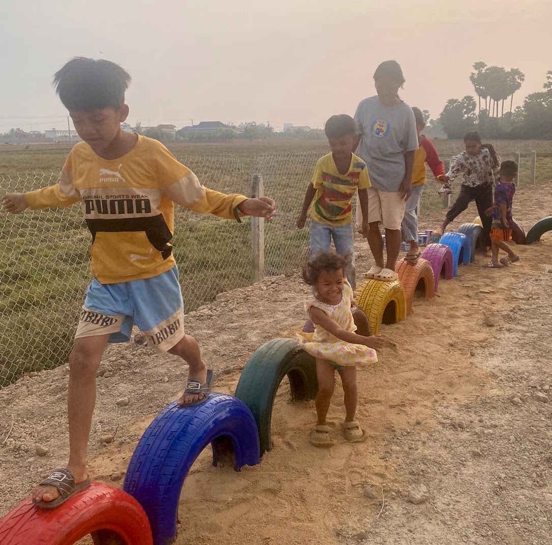 Children playing on a tire playground at dusk, golden haze over the rice fields of Pursat